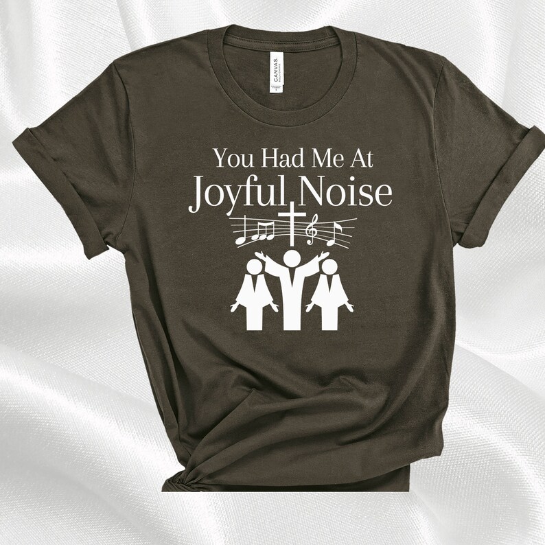 May include: A dark brown t-shirt with the text "You Had Me At Joyful Noise" in white. The text is accompanied by a graphic of three people singing with musical notes and a cross.