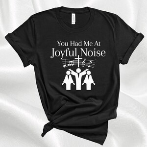 May include: Black t-shirt with white text that reads "You Had Me At Joyful Noise". The text is accompanied by a graphic of three people singing with musical notes and a cross above them.