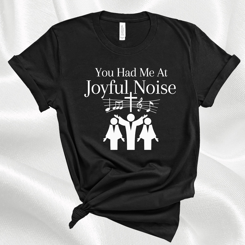 May include: Black t-shirt with white text that reads "You Had Me At Joyful Noise". The text is accompanied by a graphic of three people singing with musical notes and a cross above them.