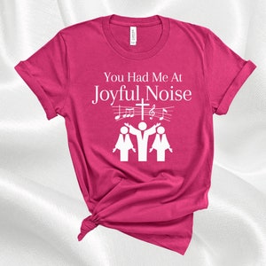 May include: Pink t-shirt with white text that reads "You Had Me At Joyful Noise". The text is accompanied by a graphic of three people singing with musical notes and a cross.