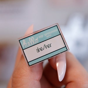 May include: A light blue name tag with the text "hello my pronouns are: she/her" in white.