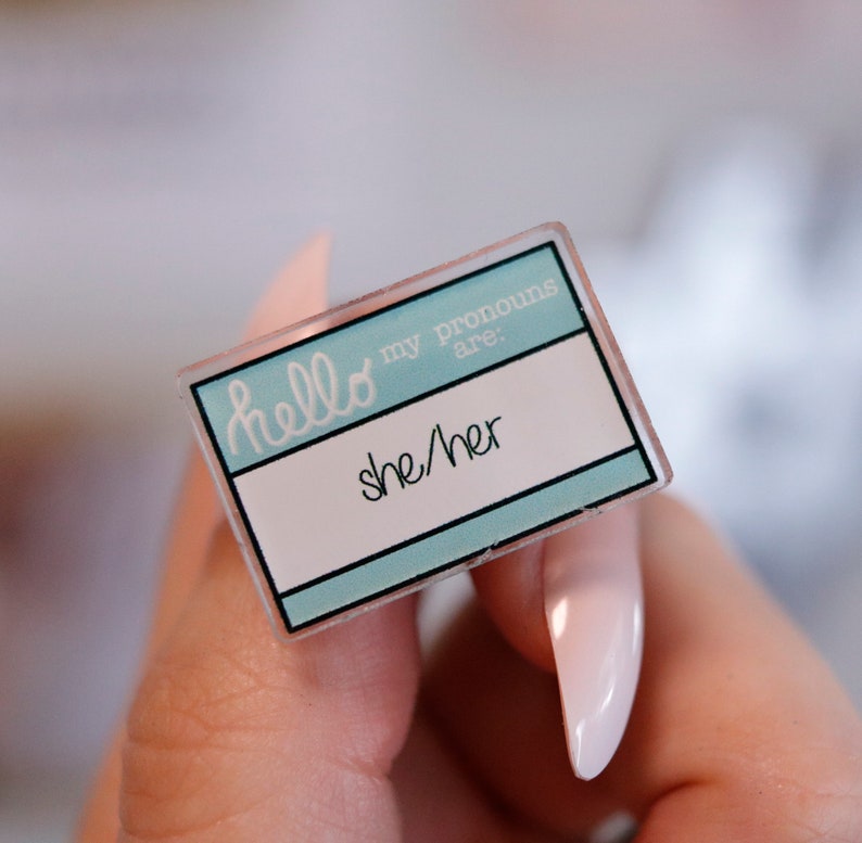 May include: A light blue name tag with the text "hello my pronouns are: she/her" in white.