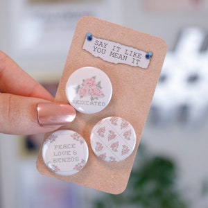 May include: Three round pinback buttons with floral designs and text. One button says "MEDICATED" with pink roses. Another says "PEACE LOVE & BENZOS" with floral accents. The third has a pink floral pattern. A small paper with "SAY IT LIKE YOU MEAN IT" is pinned above.