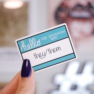 May include: A blue and white pronoun sticker with the text "Hello my pronouns are: they/them".