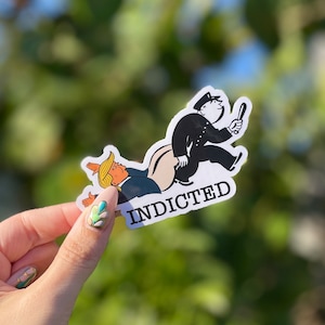 May include: A white sticker with a black and white illustration of a police officer chasing a person with the word "INDICTED" printed in black.