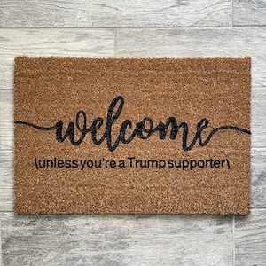 May include: A brown coir doormat with the word "welcome" written in black cursive script. Below the word "welcome" is the text "(unless you're a Trump supporter)".