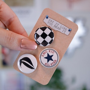 May include: A set of four button pins featuring a black and white checkered design, a blue star on a white circle with the text "Emo Kid All Star", a black silhouette of a side ponytail, and a piece of paper with the text "Say it like you mean it".