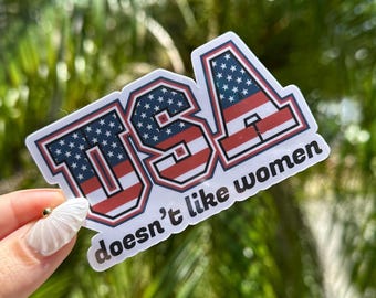 Può includere: Un adesivo bianco con la scritta "USA" in rosso, bianco e blu, con un disegno della bandiera americana. Il testo "doesn't like women" è sotto la parola "USA".