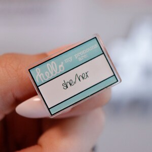 May include: A light blue plastic name tag with the text "hello my pronouns are: she/her" printed in white.