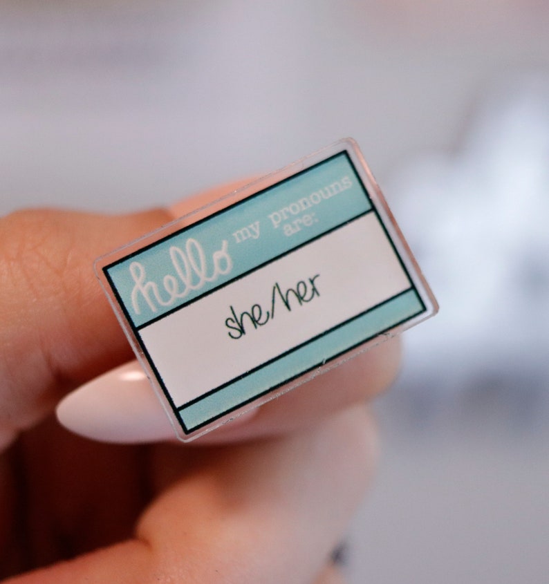 May include: A light blue plastic name tag with the text "hello my pronouns are: she/her" printed in white.