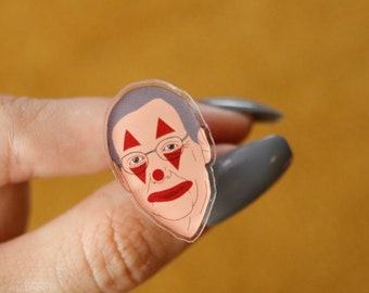Può includere: Uno spillo di plastica trasparente con un'immagine di cartone animato di una persona con trucco da clown rosso e occhiali.