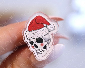 May include: A clear acrylic charm featuring a white skull wearing a red Santa hat with white trim. The skull has black eyes and rosy cheeks.