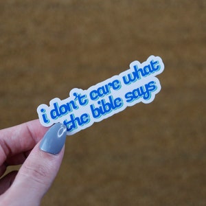 May include: A white sticker with blue text that reads "i don't care what the bible says". The text is in a retro font style.