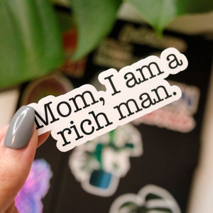 May include: A white sticker with the text "Mom, I am a rich man." in a black, bold font. The sticker is held by a person with a gray nail polish. The background is black with other stickers and a green plant.