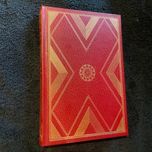 Pode incluir: Um livro de capa dura vermelho com desenhos geométricos dourados. A capa apresenta um padrão central em forma de estrela e detalhes lineares. A lombada também é vermelha, com detalhes dourados. O livro está fechado e em bom estado.