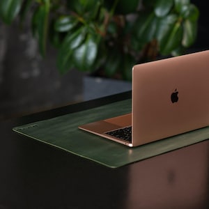 Op de afbeelding: Een roségouden laptop met een zwart toetsenbord en het Apple-logo staat open op een groene bureaumat. De laptop staat op een zwart bureau met een plant op de achtergrond. Het bureau staat in een moderne kantooromgeving.