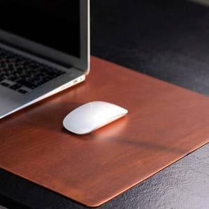 May include: A laptop and a white computer mouse sit on a copper-colored desk mat. The mat has a rectangular shape and a smooth surface. The laptop is silver and black, and the mouse is white.