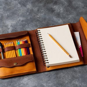 Op de afbeelding: Een bruin leren schetsboek met een ingebouwde potloodhouder. Het schetsboek is open om een blanco pagina en een geel potlood te laten zien. De potloodhouder bevat een verscheidenheid aan kleurpotloden en een zwarte pen.