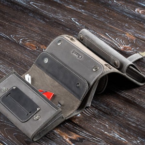 Puede incluir: Cartera de cuero gris con m&uacute;ltiples compartimentos, asegurada con broches. La cartera tiene un panel gris oscuro en contraste y el nombre "Chaus" est&aacute; grabado en la superficie. Un peque&ntilde;o art&iacute;culo rojo es visible en el interior.