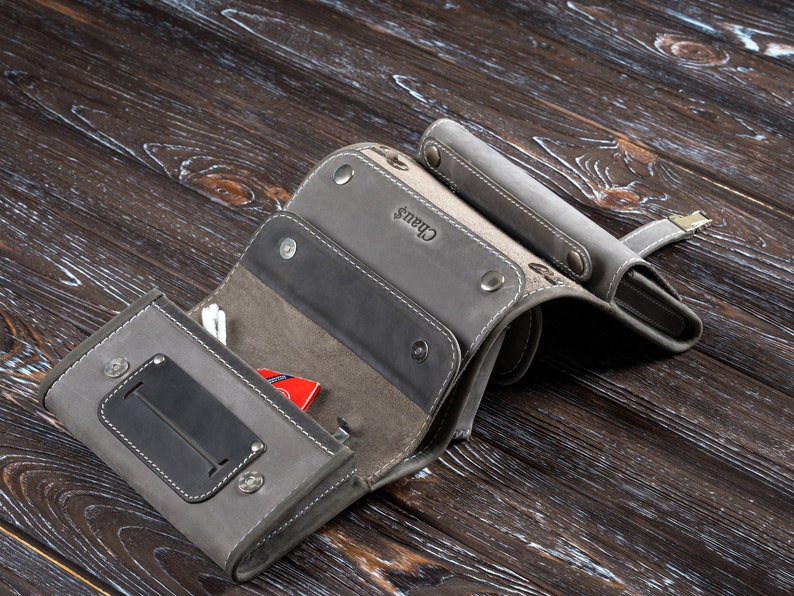 Puede incluir: Cartera de cuero gris con m&uacute;ltiples compartimentos, asegurada con broches. La cartera tiene un panel gris oscuro en contraste y el nombre "Chaus" est&aacute; grabado en la superficie. Un peque&ntilde;o art&iacute;culo rojo es visible en el interior.