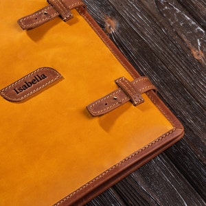May include: A yellow leather journal with brown leather straps and stitching. The journal has the name "Isabella" embossed on the front.