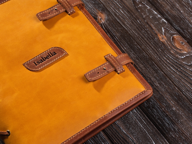 May include: A yellow leather journal with brown leather straps and stitching. The journal has the name "Isabella" embossed on the front.