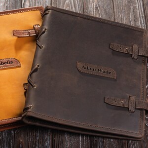 May include: Two personalized leather journals with brown and tan covers. The brown journal has the name "Adam Wade" embossed on the front. The tan journal has the name "Isabella" embossed on the front.