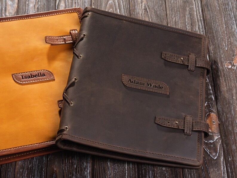 May include: Two personalized leather journals with brown and tan covers. The brown journal has the name "Adam Wade" embossed on the front. The tan journal has the name "Isabella" embossed on the front.
