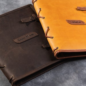 May include: Two personalized leather-bound journals with brown and tan covers. The brown journal has the name "Adam Wade" embossed on the front, and the tan journal has the name "Isabella" embossed on the front.
