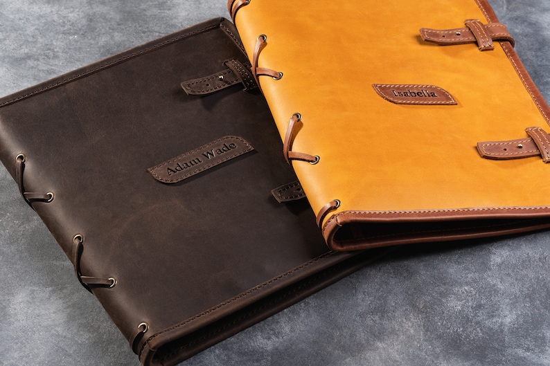 May include: Two personalized leather-bound journals with brown and tan covers. The brown journal has the name "Adam Wade" embossed on the front, and the tan journal has the name "Isabella" embossed on the front.