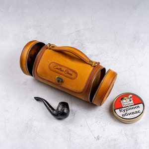 May include: A brown leather travel case with a zippered opening and a handle. The case has the words "Exelllan Chaus" embossed on the front. A black pipe and a round tin of "Devil's Holiday" tobacco are also pictured.