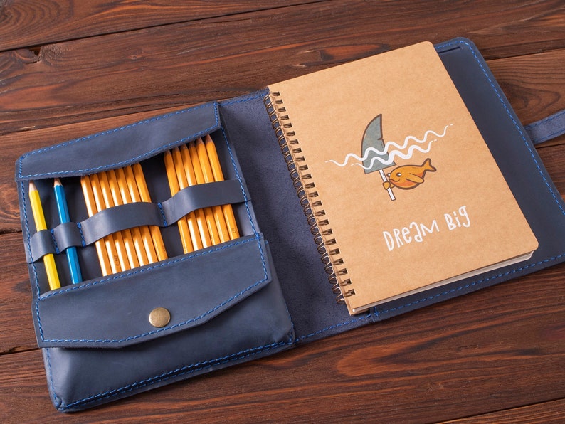 May include: A blue leather notebook with a pencil holder and a brown notebook with a shark and fish illustration and the text "Dream Big".