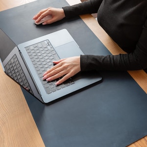 May include: A laptop computer with a silver keyboard and a black frame rests on a dark blue desk mat. The laptop is open, and a person's hands are visible, one on the keyboard and the other on a white mouse. The desk is made of wood.