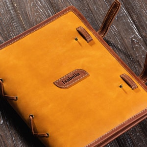 May include: A brown leather journal with a tan cover and the name "Isabella" embossed on the front. The journal has a lace-up closure and brown leather straps.