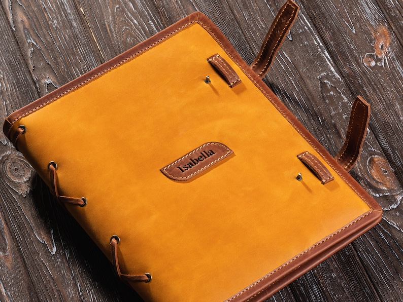 May include: A brown leather journal with a tan cover and the name "Isabella" embossed on the front. The journal has a lace-up closure and brown leather straps.
