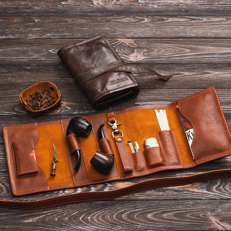 May include: A brown leather tobacco pouch with a strap, containing a pipe, a lighter, and other accessories. The pouch is open and laid flat on a wooden surface.