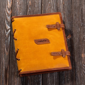 May include: A yellow leather-bound journal with brown leather accents and a brown leather strap. The journal has the name "Isabella" embossed on the front cover.