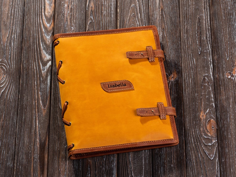 May include: A yellow leather-bound journal with brown leather accents and a brown leather strap. The journal has the name "Isabella" embossed on the front cover.