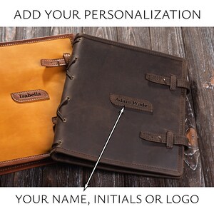 May include: Two brown leather journals with a lace-up closure. One journal has the name "Isabella" embossed on it, and the other has the name "Adam Wade" embossed on it.  The journals are on a wooden surface.