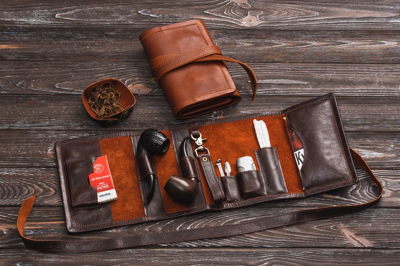 May include: A brown leather tobacco pouch with a strap, open to reveal a pipe, a pack of filters, a lighter, and other accessories.