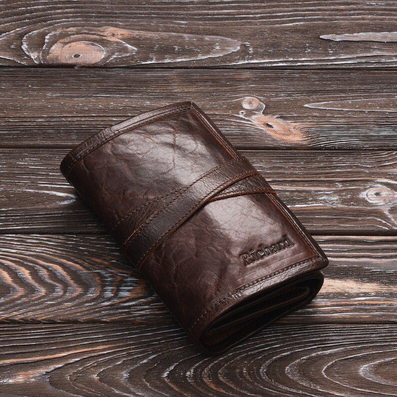May include: A brown leather wallet with a fold-over closure and a stitched strap. The wallet has the word "Richard" embossed on the front.