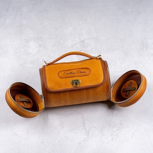 May include: A brown leather cylindrical bag with a top handle and a metal clasp. The bag has a personalized inscription that reads "Evellion Chaus".