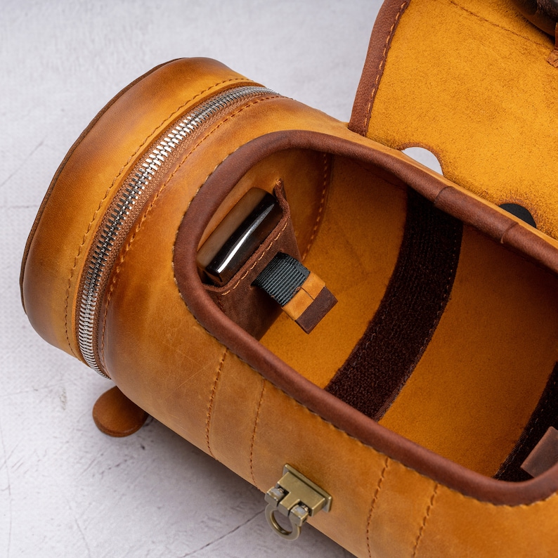 May include: A tan leather travel case with a zipper closure and a brown fabric lining. The case has a strap and a metal clasp.