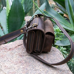 Puede incluir: Un bolso bandolera de cuero marr&oacute;n con una correa para el hombro. El bolso tiene m&uacute;ltiples compartimentos y est&aacute; adornado con herrajes y costuras doradas. El bolso est&aacute; sobre una roca con plantas verdes en el fondo.