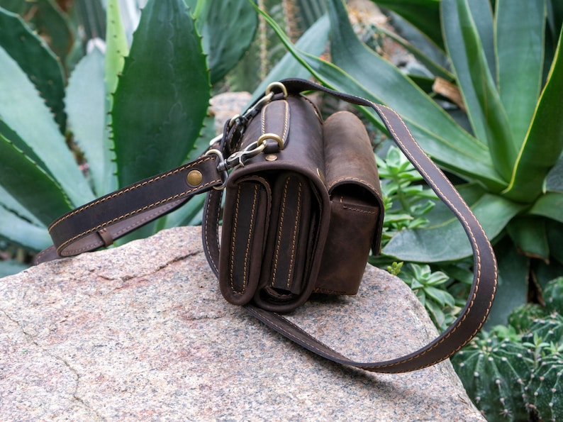 Puede incluir: Un bolso bandolera de cuero marr&oacute;n con una correa para el hombro. El bolso tiene m&uacute;ltiples compartimentos y est&aacute; adornado con herrajes y costuras doradas. El bolso est&aacute; sobre una roca con plantas verdes en el fondo.