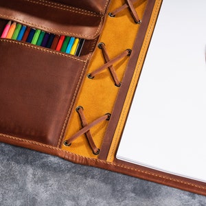 May include: A brown leather portfolio with a yellow leather flap and a lace closure. The portfolio has a pocket for pencils and a notepad with a pencil on the right side.