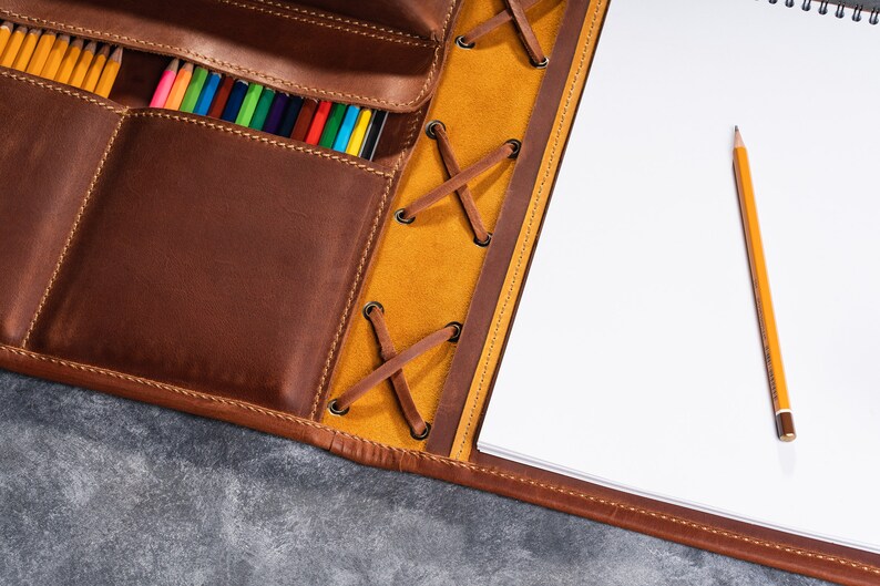 May include: A brown leather portfolio with a yellow leather flap and a lace closure. The portfolio has a pocket for pencils and a notepad with a pencil on the right side.