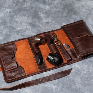 May include: A brown leather tobacco pipe roll containing a pipe, a black pipe, and a small silver tool. The roll is open, revealing a tan interior. The roll is made of brown leather and has a strap to secure it.