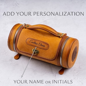 May include: A tan leather cylindrical travel bag with a top handle and a zipper closure. The bag has a small rectangular patch with the text "Evolution Chaos" embossed on it. The text "YOUR NAME OR INITIALS" is written below the bag.