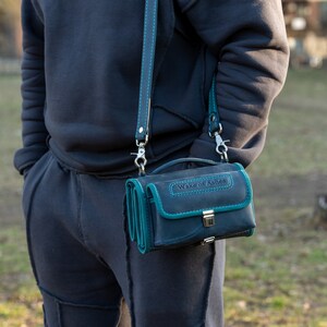 Puede incluir: Un bolso bandolera de cuero azul oscuro con ribetes y costuras turquesas. El bolso tiene un cierre de solapa y una correa desmontable. El texto "Wake of Ashes" est&aacute; grabado en la parte superior.